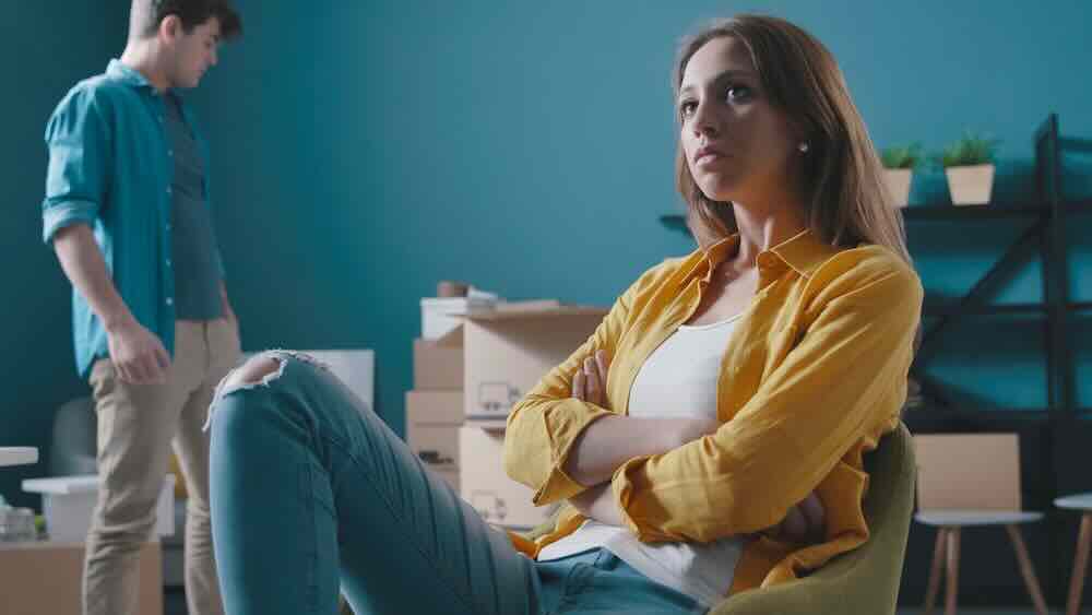 Young couple unpacks boxes and looks frustrated to tell boyfriend about pregnancy.