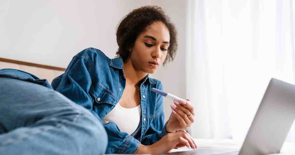 Young woman reading a pregnancy test while using her laptop in bed at home, wondering how to take a pregnancy test.