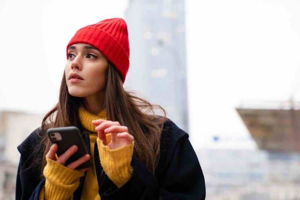 Beautiful girl with a red stocking cap standing outside researching “where is the easiest place to get tested for STDs?” on her phone